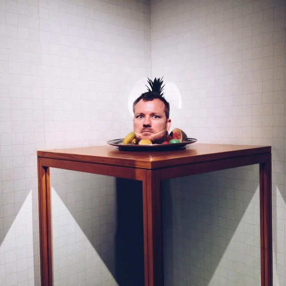 Van's head sitting on a platter of fruit with a pineapple top for hair, on a timber side table in a papered corner