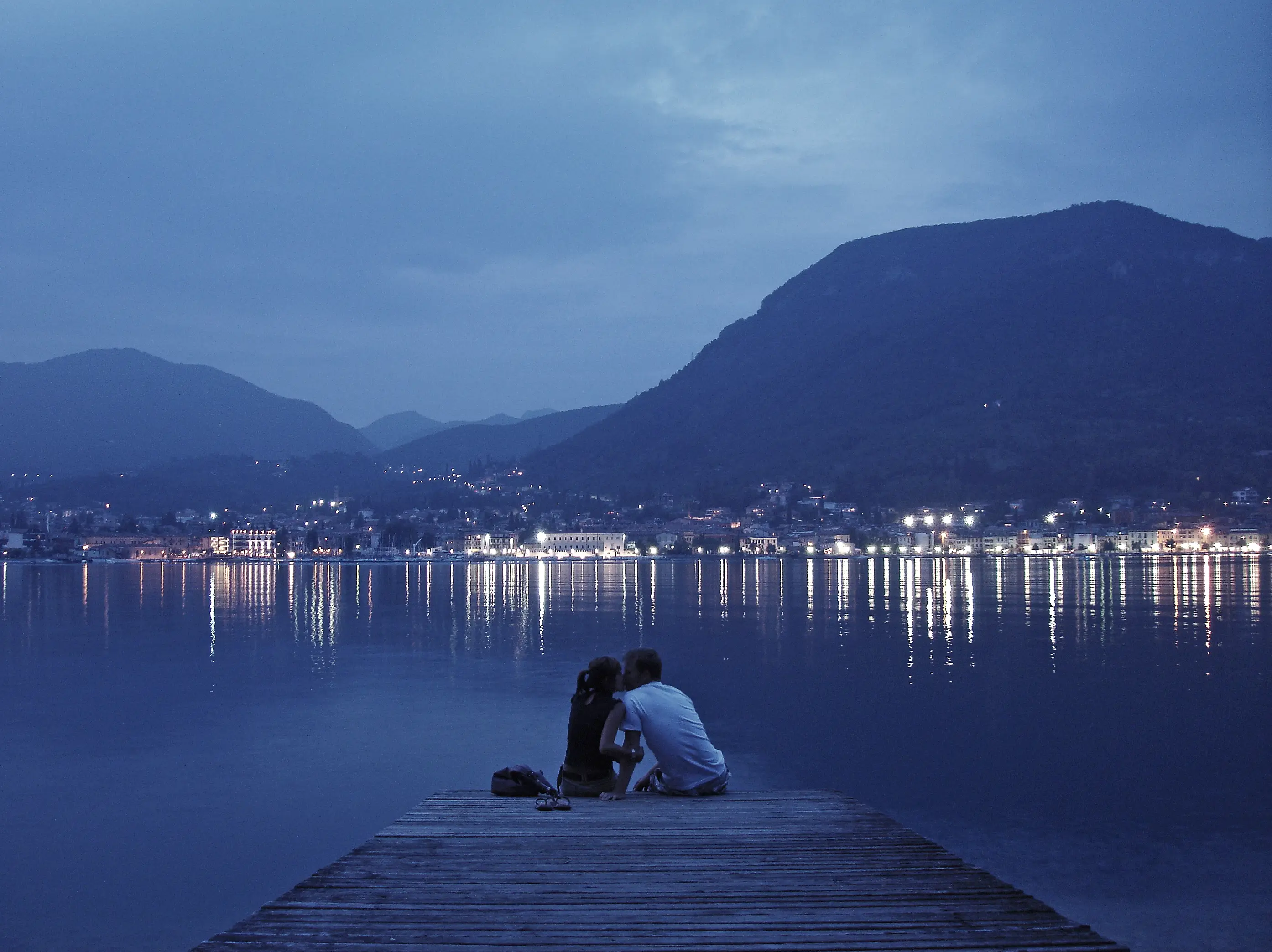 A couple kissing at the end of a timber jetty at blue hour, town lights reflecting across a lake below dark mountains