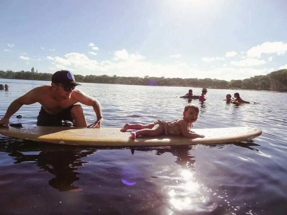 Van in the water beside a longboard with his baby daughter lying on it, swimmers in the lake behind